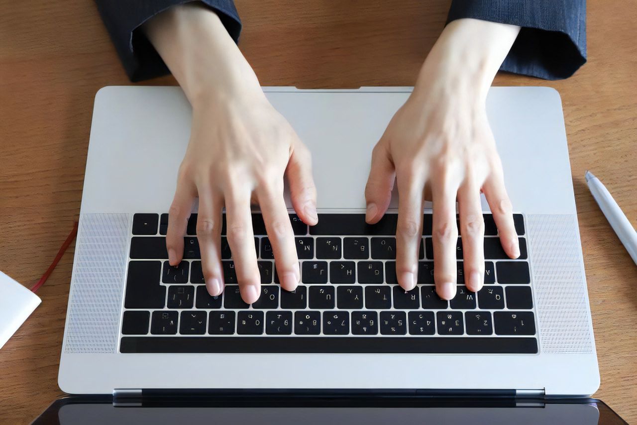 木の机に置いたノートパソコンのキーボードに両手を添えて操作している様子を真上から見たところ